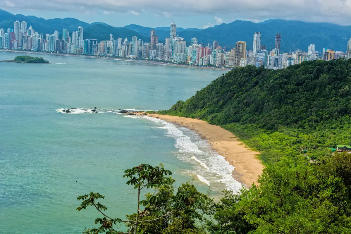 Vista aérea de praia de águas verdes e mata atlântica com os prédios de Balneário Camboriú ao fundo